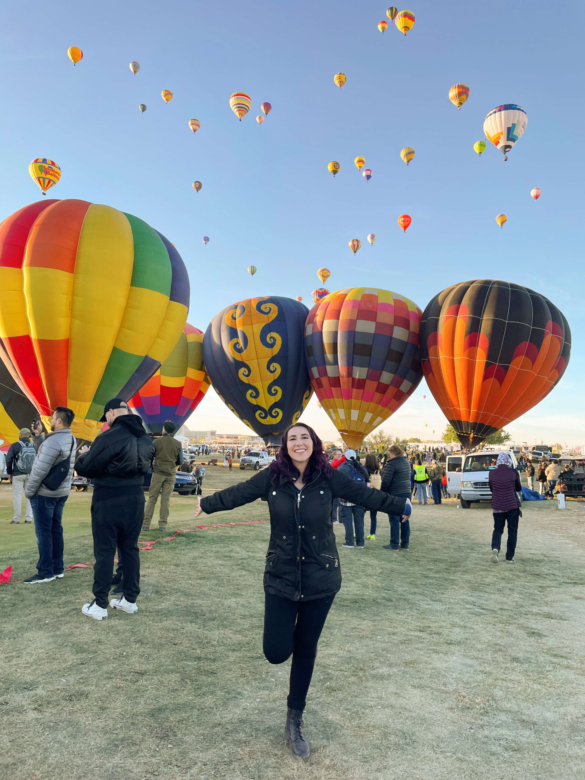 A girl smiles at the camera with her arms up in the air in front of 4 big hot air balloons at the balloon fiesta, with many small hot air balloons in the sky above her