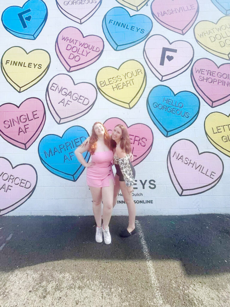 two friends smiling in front of heart shaped mural in nashville