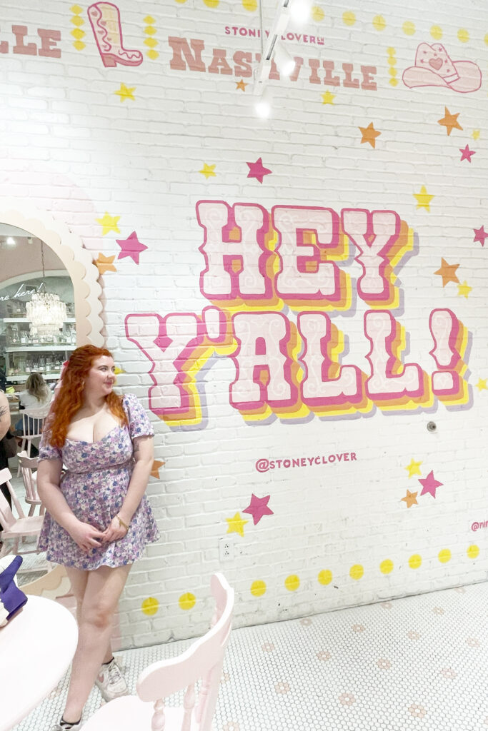 girl in floral dress, smiling in front of a mural that says hey yall in nashville