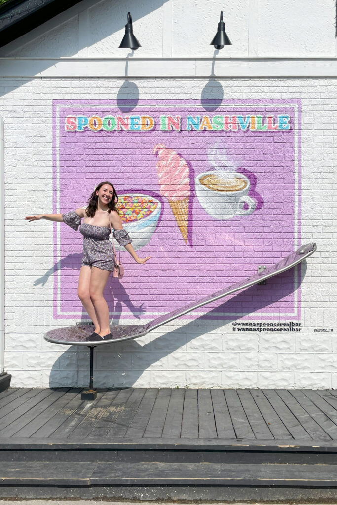 girl smiling while standing on an oversized spoon installation with a mural that says spooned nashville